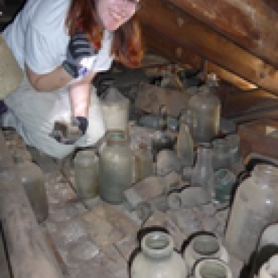 woman with a flashlight crouching in an attic full of glassware