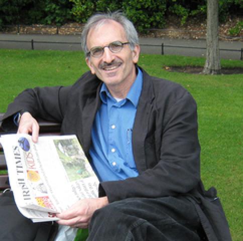 man in a blue shirt and charcoal sport coat reading a newspaper on a bench