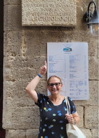 Woman in a black patterned shirt smiling and pointing upwards to a sign from the University of Catania Archaeology Institute